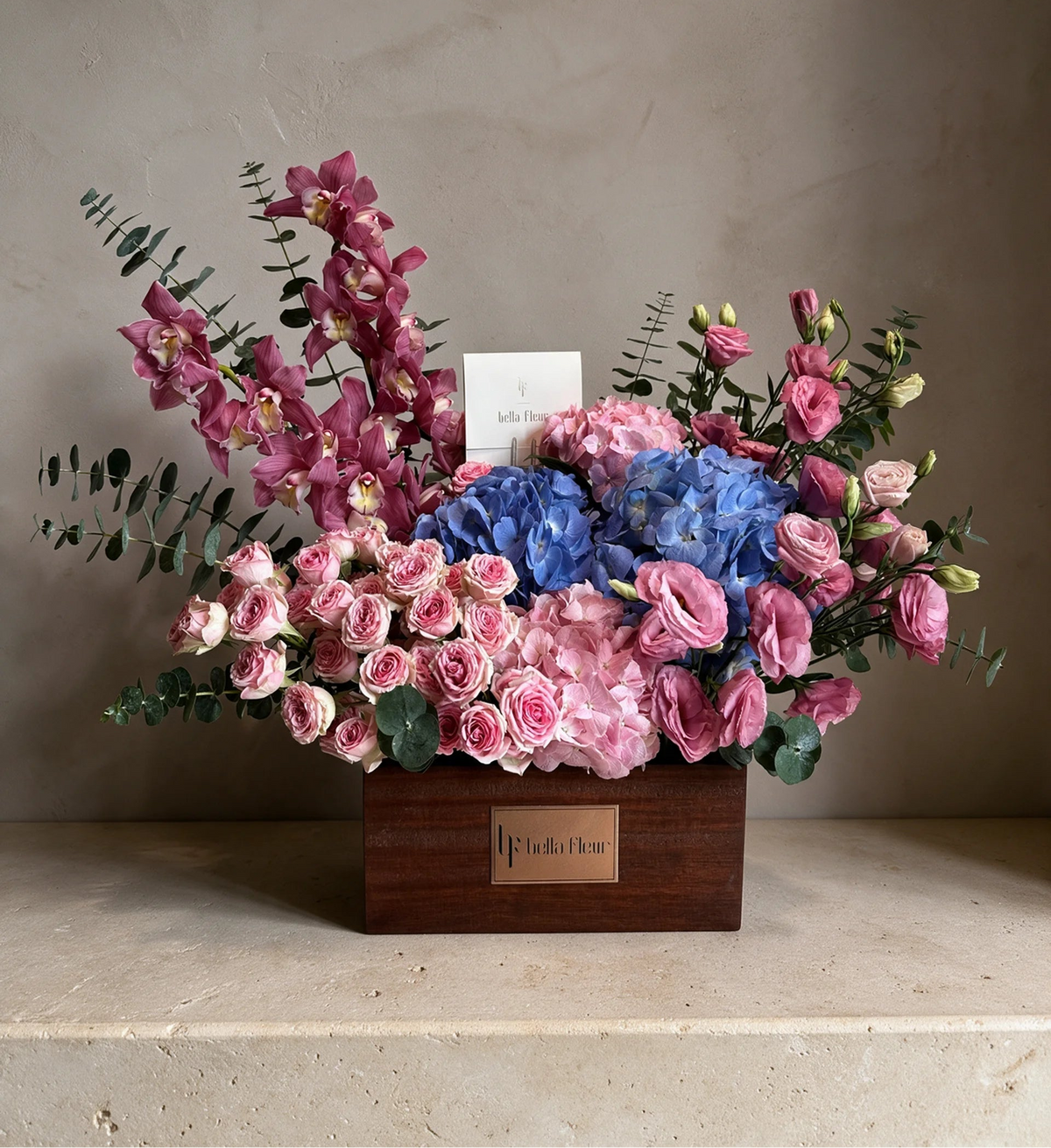 A floral arrangement in a rectangular wooden box on a stone surface against a textured beige wall. The bouquet features pink roses, blue hydrangeas, pink orchids, pink lisianthus, green eucalyptus, and a small white card.